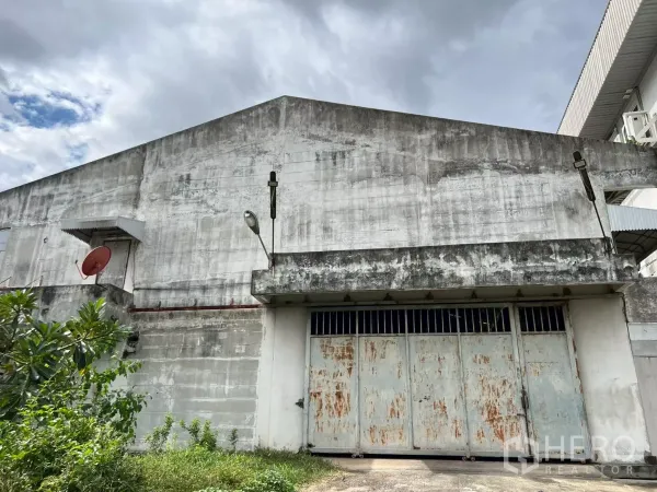 คลังสินค้า ให้เช่าในPhra Khanong, Bangkok - ด้านหน้าโกดังอุตสาหกรรม แสดงประตูบานเลื่อนขนาดใหญ่ ไฟส่องสว่าง และจานดาวเทียม บนผนังคอนกรีตภายใต้ท้องฟ้าครึ้ม