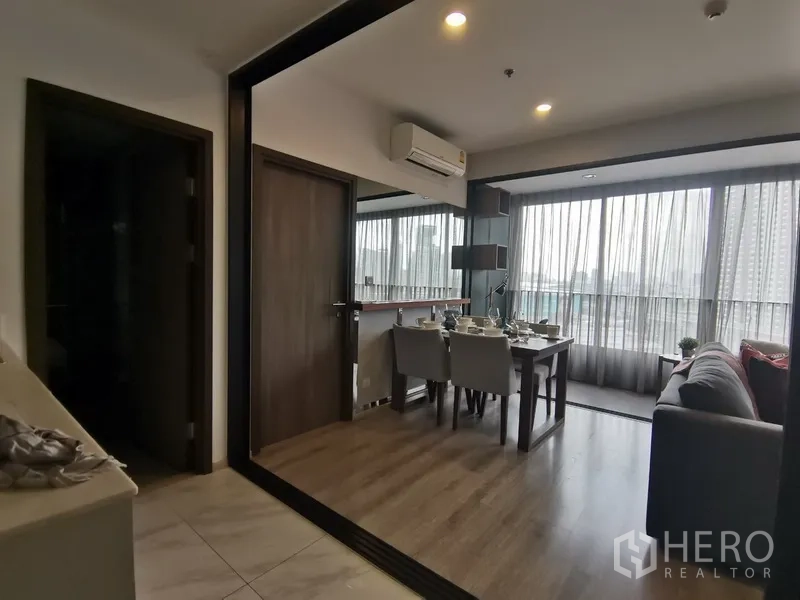 Condominium for rent in Huai Khwang, Bangkok - Dining area and lounge opening to a glass‑enclosed balcony with sheer curtains and warm wood flooring.