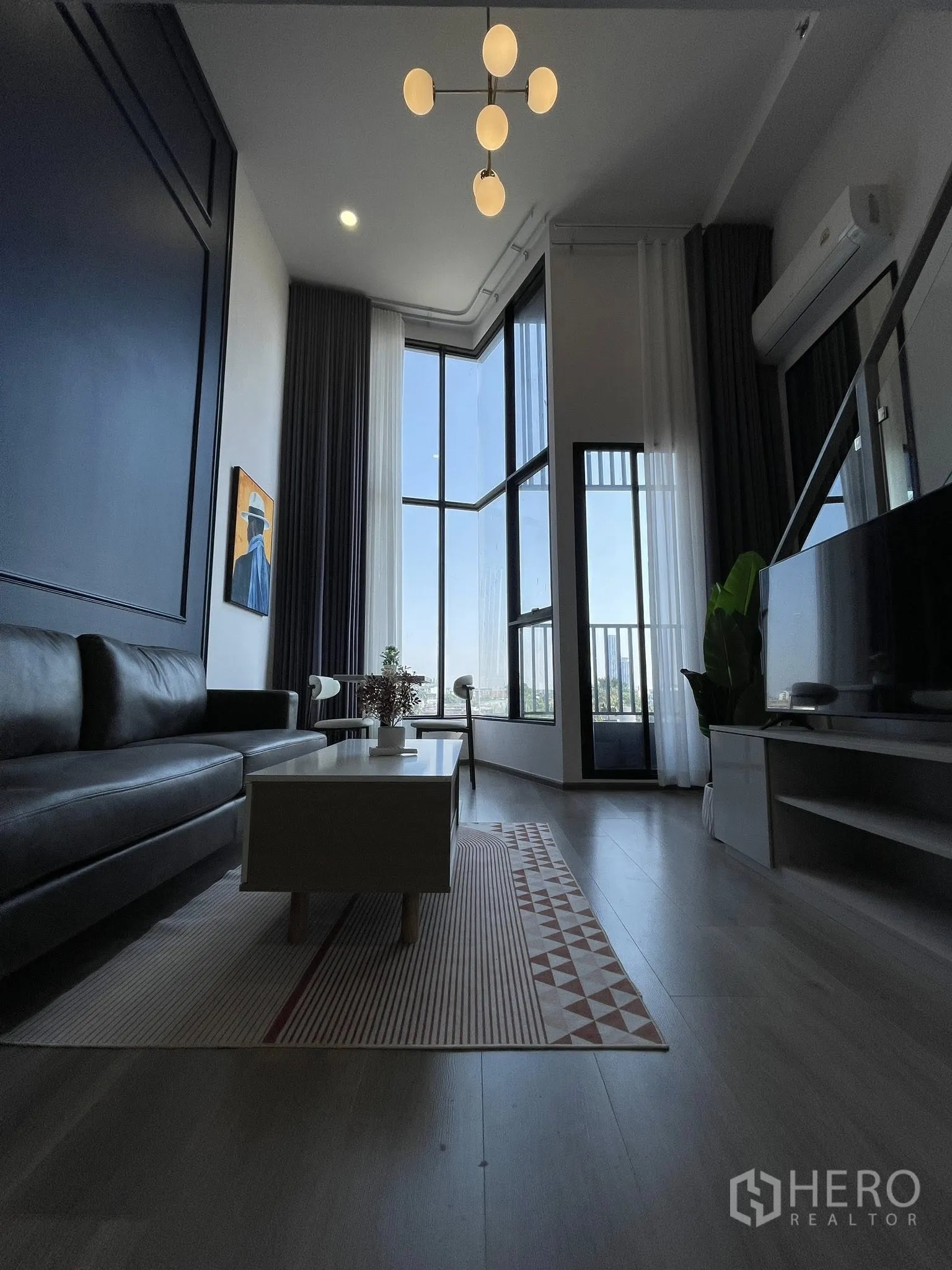 Condominium for rent in Khlong Toei, Bangkok - Loft-style living room with high ceiling, chandelier, sofa, TV console, and large windows.