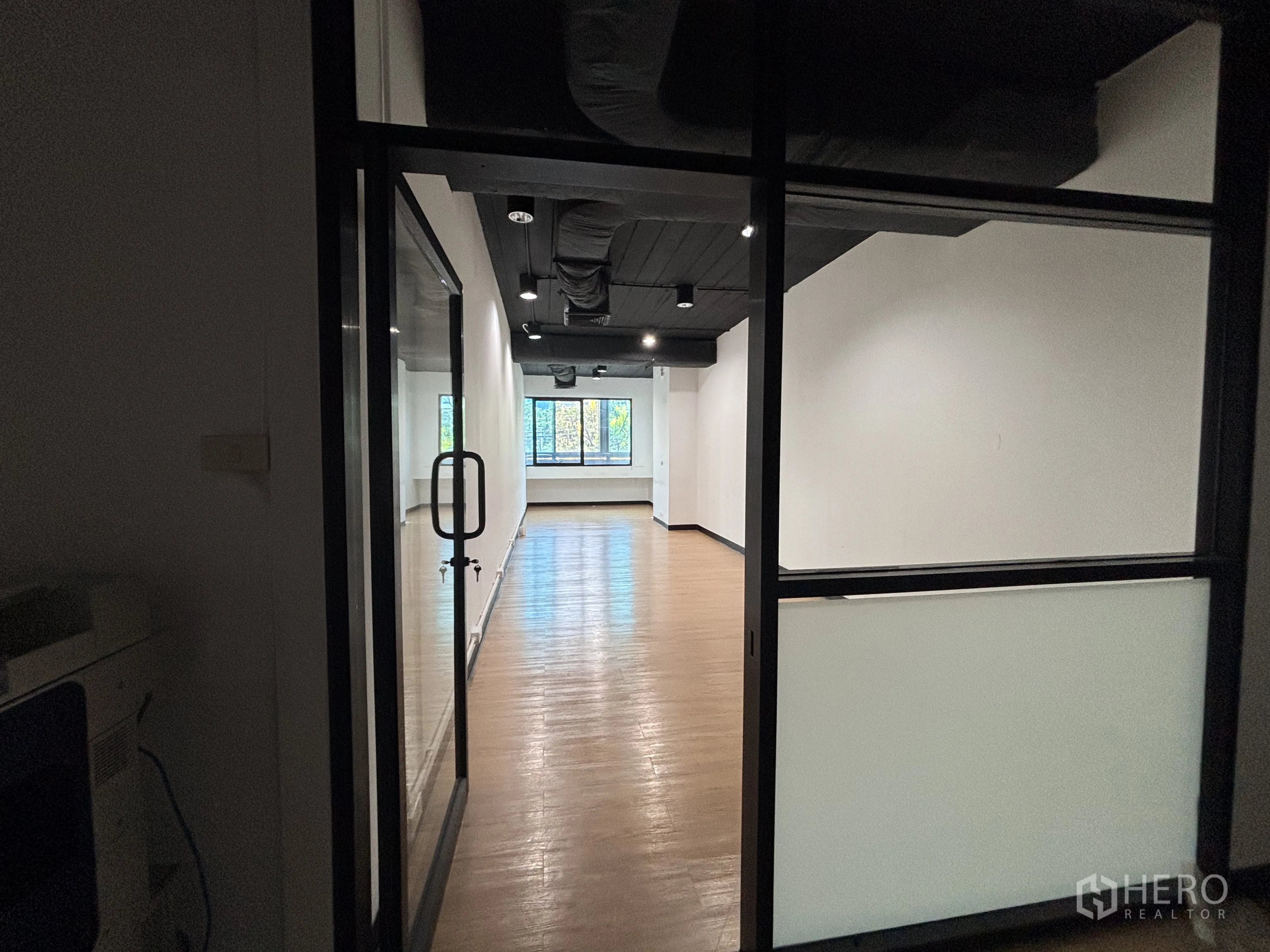 Office Space for rent in Bang Rak, Bangkok - Long room behind a glass door with wood floors, black ceiling, and windows at the far end.