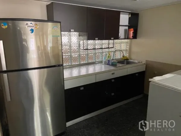 Townhouse for rent in Khlong Toei, Bangkok - Compact fitted kitchen with stainless-steel refrigerator, glass-block window, sink, and cabinetry.