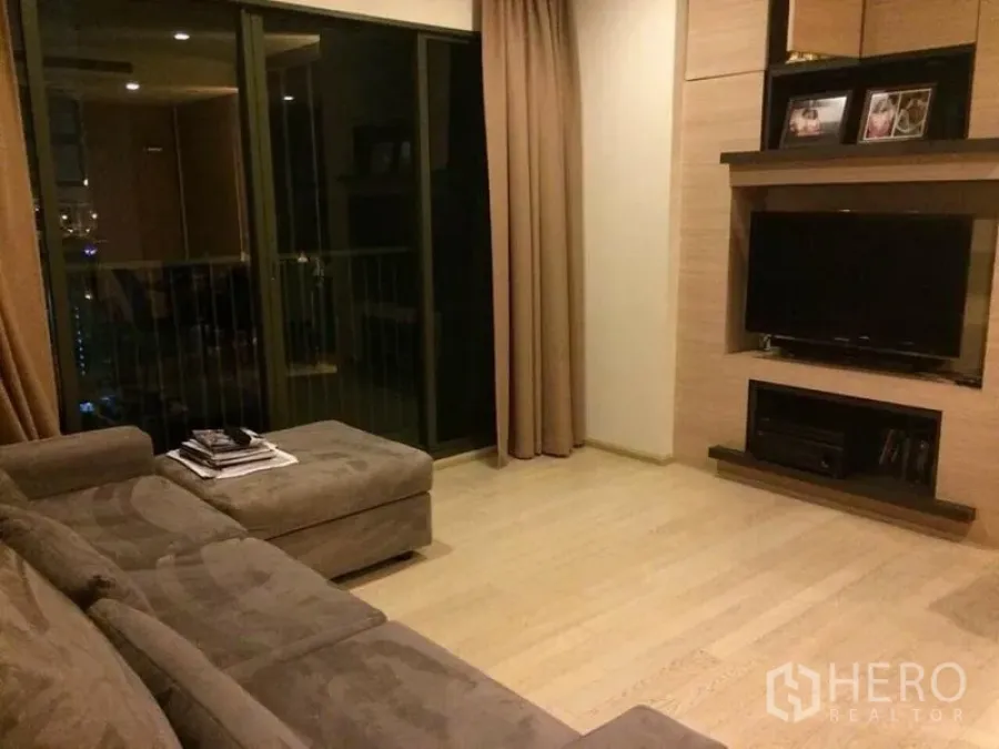 Condominium for rent in Watthana, Bangkok - Living room with sectional sofa, TV wall unit, and sliding glass door to a balcony.