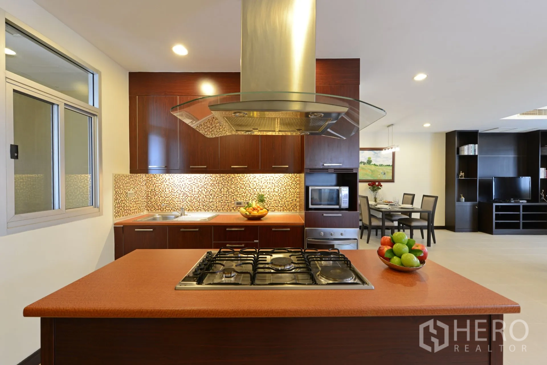 Apartment for rent in Watthana, Bangkok - Kitchen island with gas hob facing the dining area and built-in shelving under warm lighting.