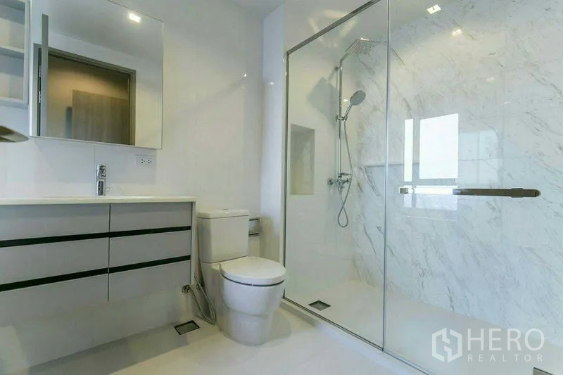 Condominium for rent or sale in Watthana, Bangkok - Bright bathroom with glass-enclosed rain shower, marble wall tiles, floating vanity and toilet.