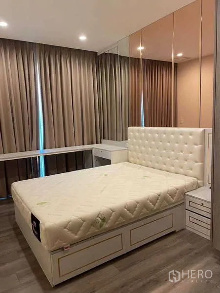 คอนโด ให้เช่า - มุมมองอีกด้านของห้องนอนหลัก เตียงควีนพร้อมลิ้นชักข้างเตียง และผ้าม่านสูงเต็มผนังกับผนังหัวเตียงกระจก