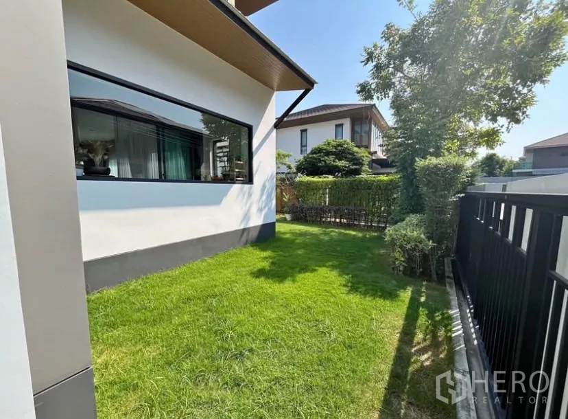 Detached House for rent in Prawet, Bangkok - Side garden with grass, black fence and a wide horizontal window of the house.