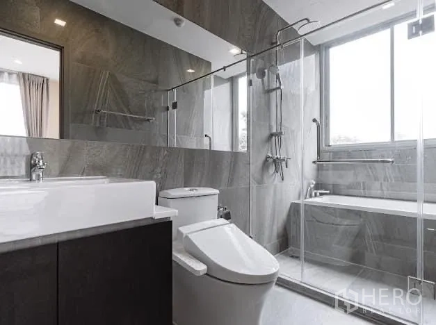 Condominium for rent in Watthana, Bangkok - Modern bathroom with glass shower, bathtub by window, and gray stone tiles.