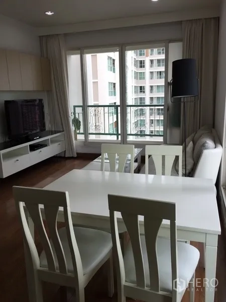 Condominium for rent in Pathum Wan, Bangkok - Dining area with a white table set beside the balcony and TV unit in a light-filled living room.