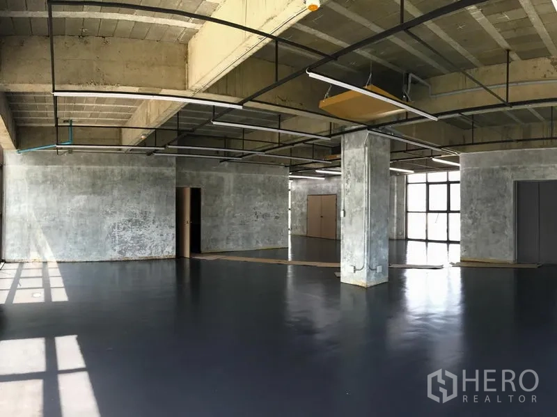 Office Space for rent in Khlong Toei, Bangkok - Industrial-style workspace with concrete columns, black epoxy floor, suspended lighting, multiple doorways, and bright corner windows.