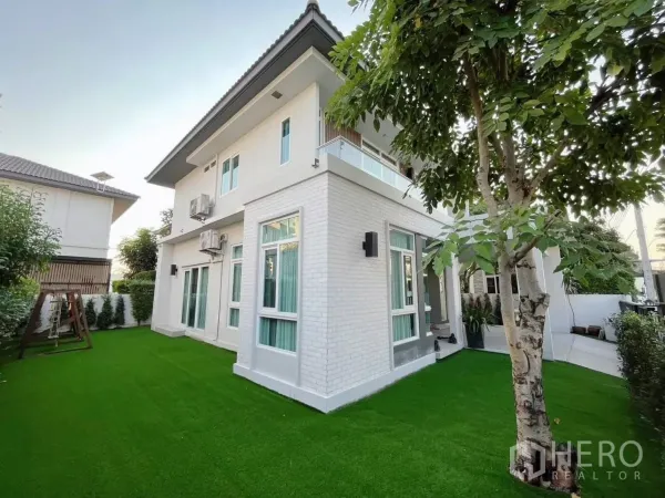 Detached House for rent in Samut Prakan - Two-storey white detached house with large green lawn and mature trees in a gated community.