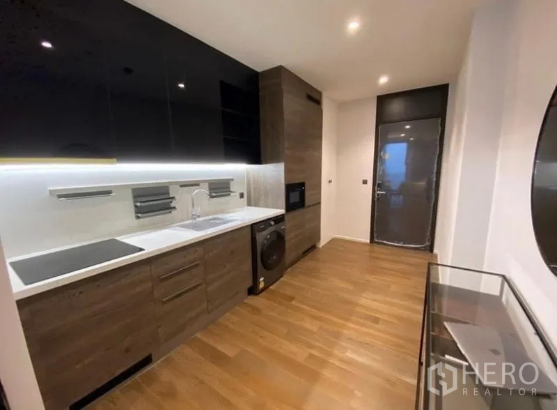 Condominium for rent in Pathum Wan, Bangkok - Sleek linear kitchen with dark cabinetry, under-cabinet lighting, sink, cooktop, built-in appliances, and glass door.