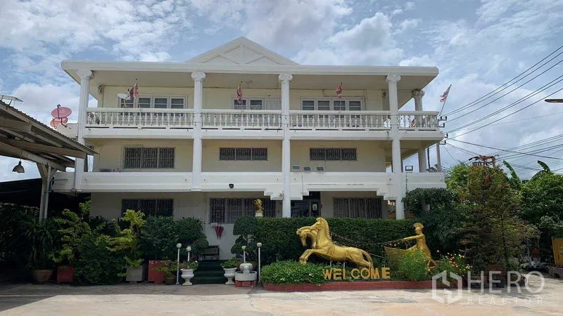 Warehouse for rent in Phra Pradaeng, Samut Prakan - Three-storey office building with balconies facing a concrete yard and garden statues.