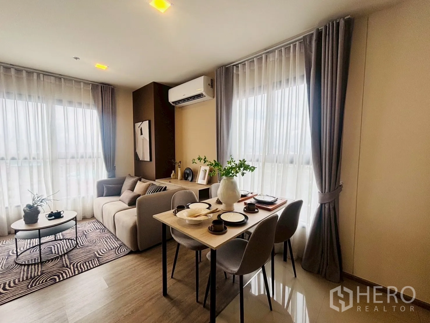 Condominium for rent in Watthana, Bangkok - Corner living and dining space with sofa, four-seat dining table, and two large windows with curtains and air conditioner.