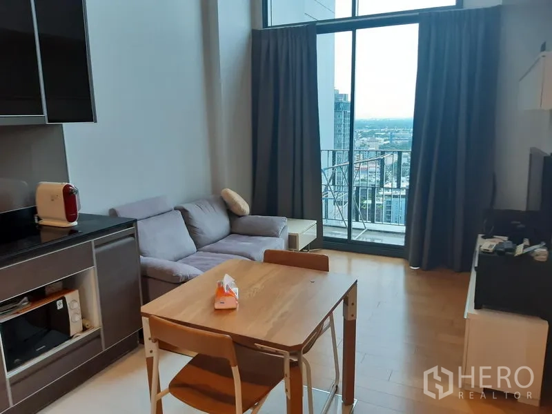 Condominium for rent or sale in Khlong Toei, Bangkok - Open‑plan living area with sofa and dining set, floor‑to‑ceiling glass doors to a balcony and wide Bangkok skyline view.