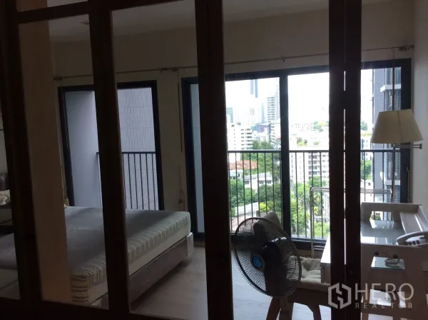 公寓 出租在Khlong Toei, Bangkok - 卧室带推拉玻璃门，连接阳台，远眺城市与绿景，旁有书桌与风扇。