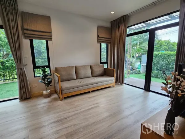 Detached House for rent or sale in Don Mueang, Bangkok - Sunlit lounge with sofa, corner windows, and a lawn view through glass doors.