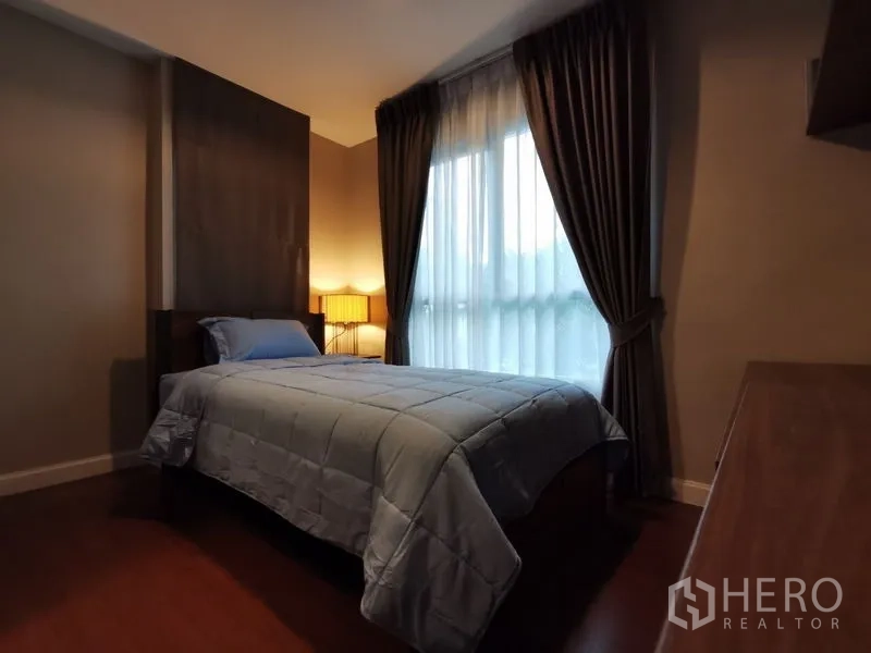 Condominium for rent or sale in Huai Khwang, Bangkok - Cozy single bedroom with gray bedding, bedside lamp, and large curtained window.