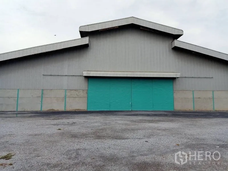 Factory for rent in Chaloem Phra Kiat, Saraburi - Industrial factory exterior with corrugated metal cladding, teal sliding doors, and a large paved yard.