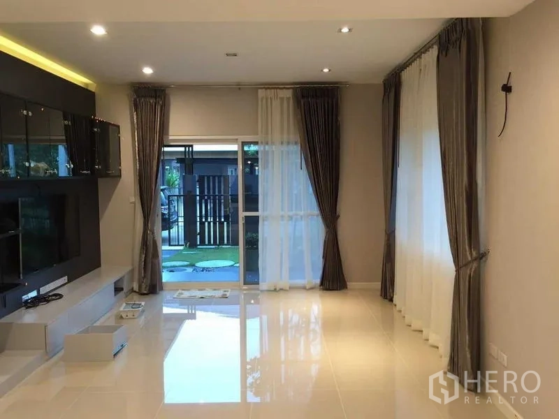 Townhouse for sale in Bang Khae, Bangkok - Light-filled living room with media wall, full-length curtains and sliding door to the garden.