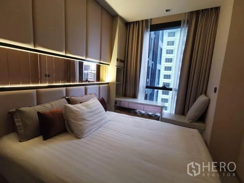 คอนโด ให้เช่าหรือขายในRatchathewi, Bangkok - ห้องนอนอบอุ่นพร้อมหัวเตียงบุผ้า หน้าต่างชมวิวเมือง และแสงไฟโทนอุ่นในคอนโดราชเทวี