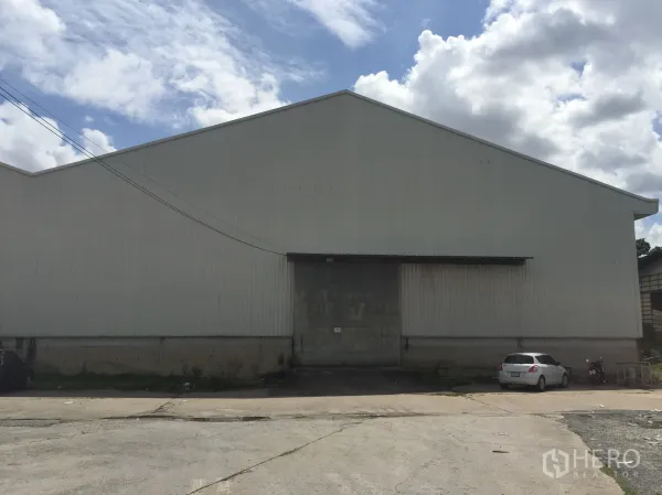 Warehouse for rent in Pak Kret, Nonthaburi - Large corrugated metal warehouse in Pak Kret, Nonthaburi featuring a high gable roof, wide loading door, and concrete driveway.