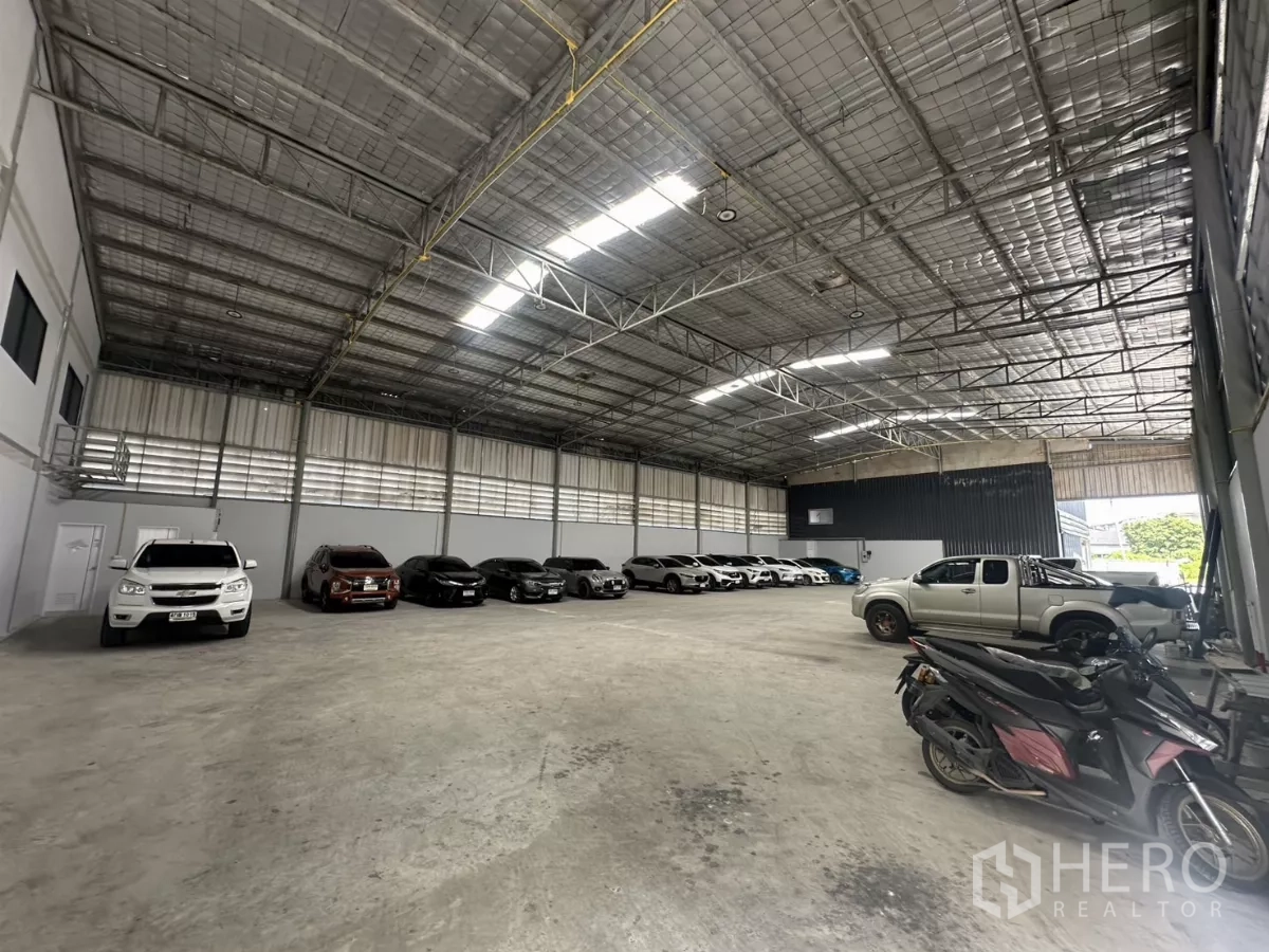 Warehouse for rent in Suan Luang, Bangkok - Open warehouse interior showing steel truss roof, roller shutter door, and a small wall-side office area.