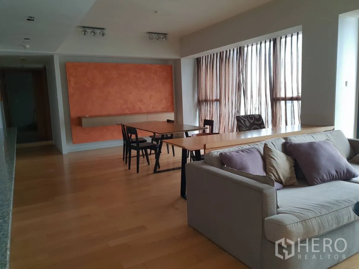 Condominium for rent in Sathon, Bangkok - Open-plan living and dining area with hardwood floors, an orange feature wall, long table, sofa and large windows with sheer curtains.