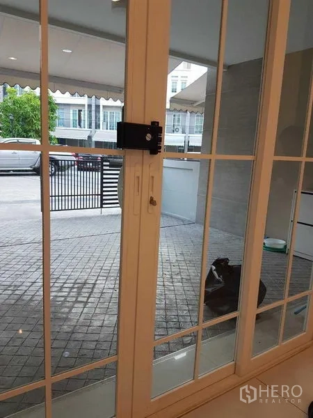 联排别墅 出租在Bang Na, Bangkok - 通过白框推拉门望向带门车棚与铺砖前庭。