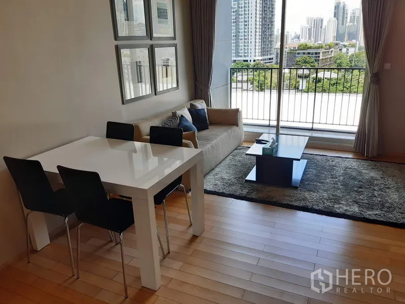 Condominium for rent in Khlong Toei, Bangkok - Bright living and dining area with sofa, table, floor-to-ceiling window and balcony overlooking city skyline and greenery.