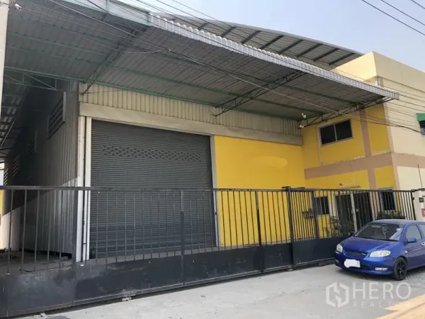 โรงงาน ให้เช่าในPathum Thani - ด้านหน้าโรงงานสีเหลือง ประตูม้วนขนาดใหญ่ หลังคาคลุมลานโหลด รั้วเหล็ก และรถสีน้ำเงินจอดหน้าทางเข้า