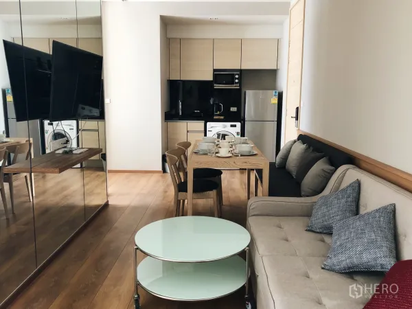 Condominium for rent in Khlong Toei, Bangkok - Open-plan living room with mirror wall, round glass coffee table and a compact kitchen beyond.