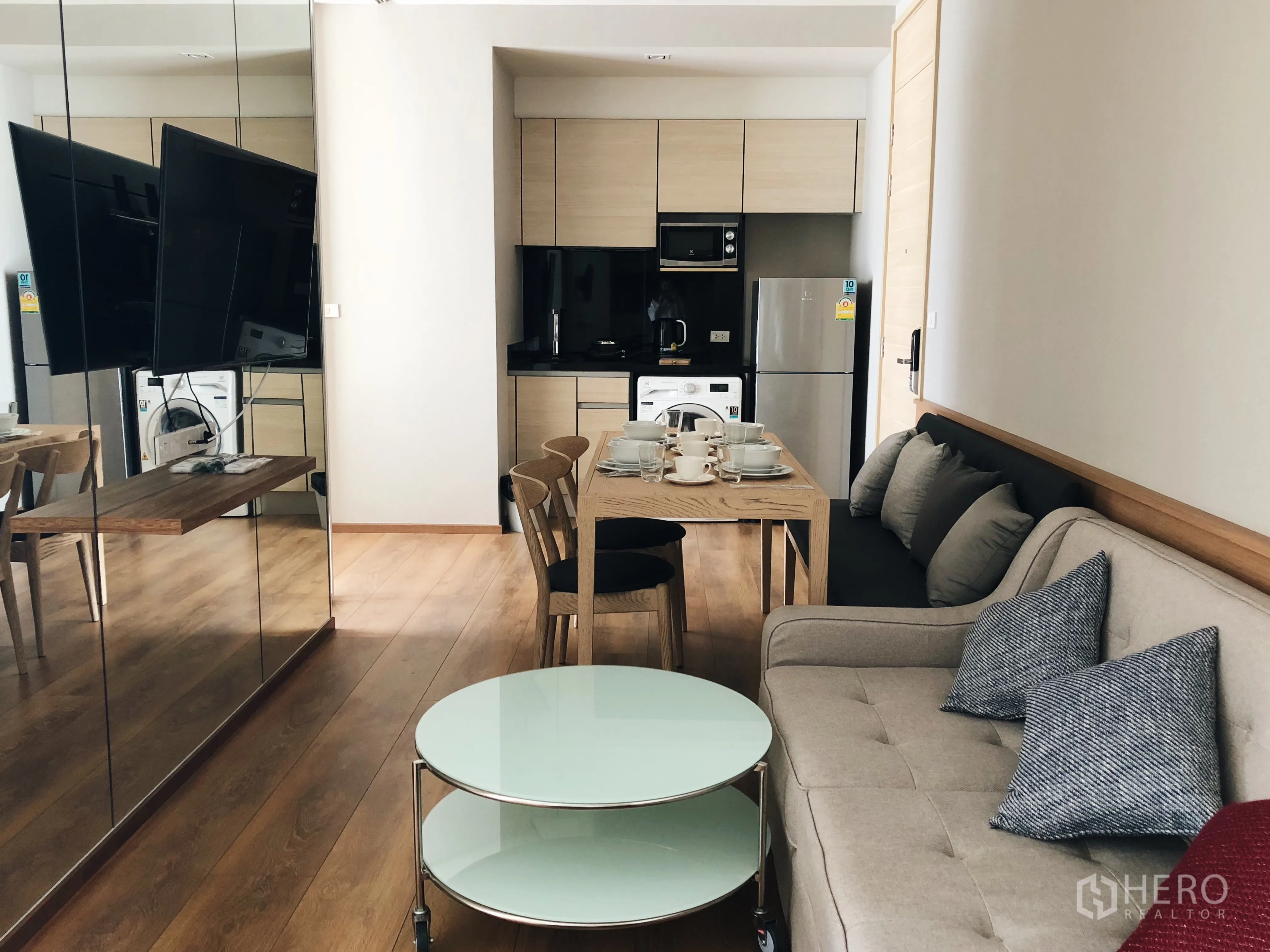 Condominium for rent in Khlong Toei, Bangkok - Open-plan living room with mirror wall, round glass coffee table and a compact kitchen beyond.
