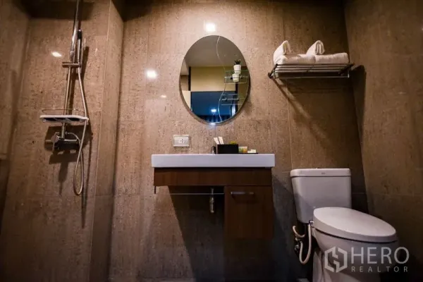 Condominium for rent in Khlong Toei, Bangkok - Contemporary bathroom featuring wall-mounted vanity, oval mirror, toilet and walk-in shower.