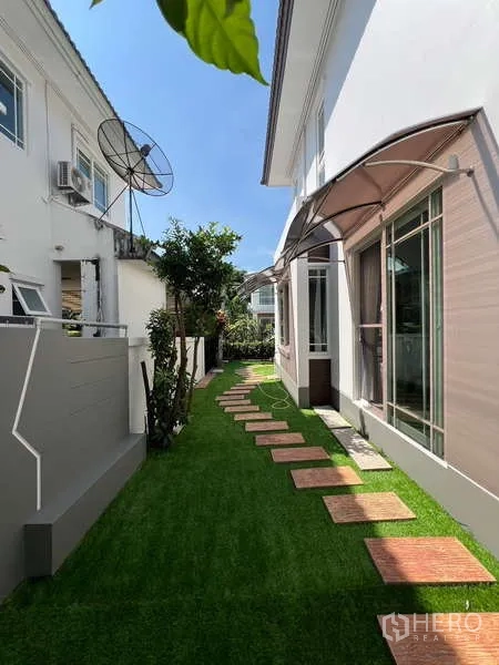 บ้าน ให้เช่าในBang Kapi, Bangkok - ทางเดินสวนข้างบ้านปูแผ่นทางเท้าบนสนามหญ้า ติดกับผนังบ้านโมเดิร์นสีขาว
