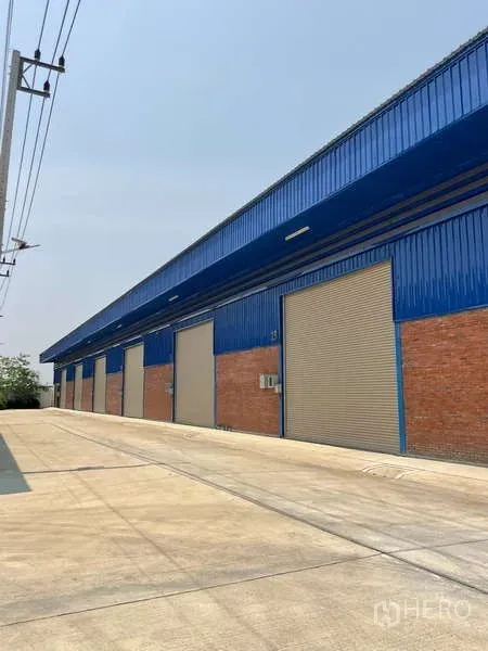 Warehouse for rent in Bang Bua Thong, Nonthaburi - Row of blue steel warehouses with brick base and multiple roller shutter doors along a wide concrete driveway.
