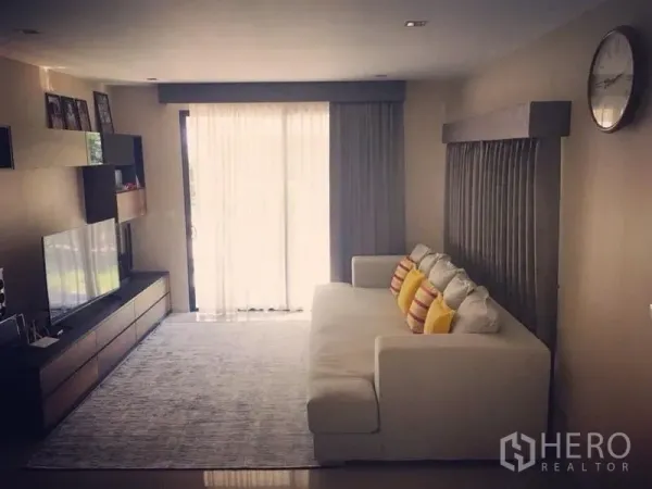 บ้าน ขาย - ห้องนั่งเล่นสว่าง โซฟาสีขาว ผนังทีวี และประตูกระจกบานเลื่อนพร้อมม่านโปร่ง