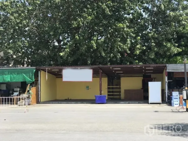 零售空间 出租在Bang Rak, Bangkok - 开放式临街铺面，黄色墙体与金属雨棚，门面宽阔，前方硬化空地被大树遮荫。