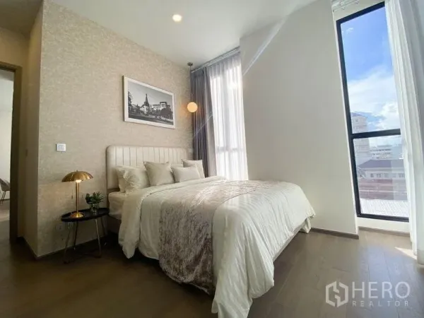 Condominium for rent in Ratchathewi, Bangkok - Primary bedroom with large windows, city view, soft lighting and a neatly made bed with neutral tones.