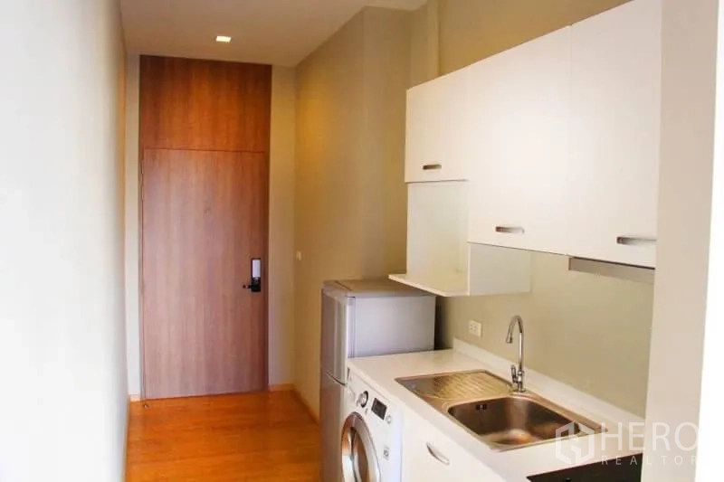 Condominium for rent or sale in Watthana, Bangkok - Galley kitchenette with white cabinets, sink, fridge and washing machine.