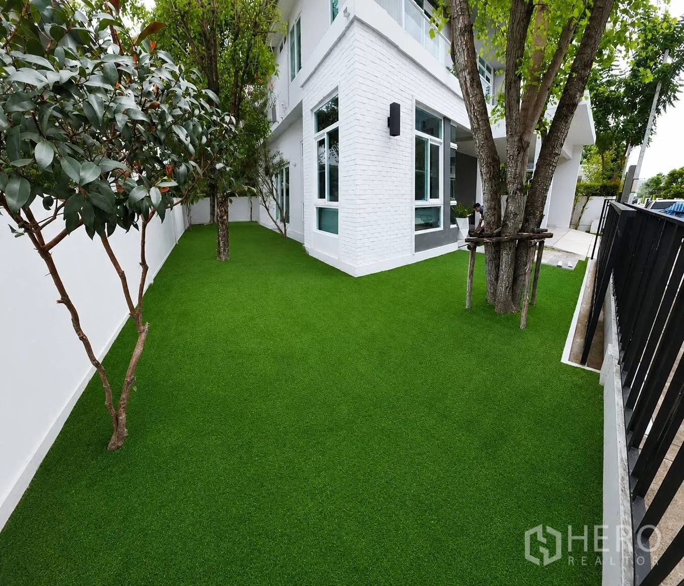 บ้านเดี่ยว for rent in Bang Phli, Samut Prakan - สวนข้างบ้านปูหญ้าเทียมพร้อมต้นไม้ใหญ่และผนังอิฐสีขาวของตัวบ้าน