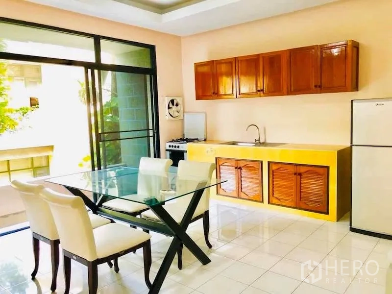 Townhouse for rent in Watthana, Bangkok - Light kitchen with wooden cabinets, glass dining table, fridge and balcony-style sliding doors.