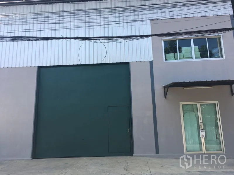 คลังสินค้า ให้เช่าในBang Kapi, Bangkok - ด้านหน้าโกดังสมัยใหม่พร้อมประตูม้วนสีเขียวขนาดใหญ่และทางเข้าออฟฟิศกระจก.