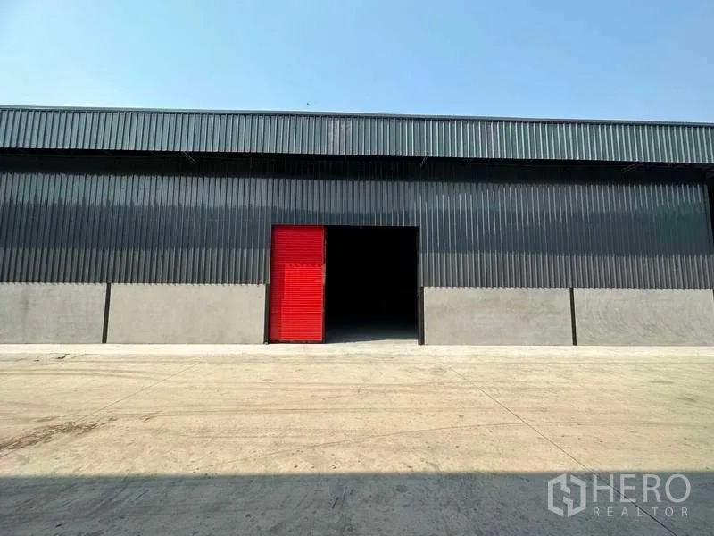 คลังสินค้า ให้เช่าในKhlong Sam Wa, Bangkok - ด้านหน้าโกดังผนังเมทัลชีทสีดำ ประตูม้วนสีแดง ลานคอนกรีตกว้าง