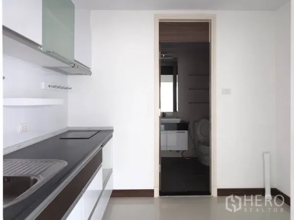 Condominium for rent in Yan Nawa, Bangkok - Minimal white kitchen with built-in hob, sink and cabinets, connected to a bathroom.