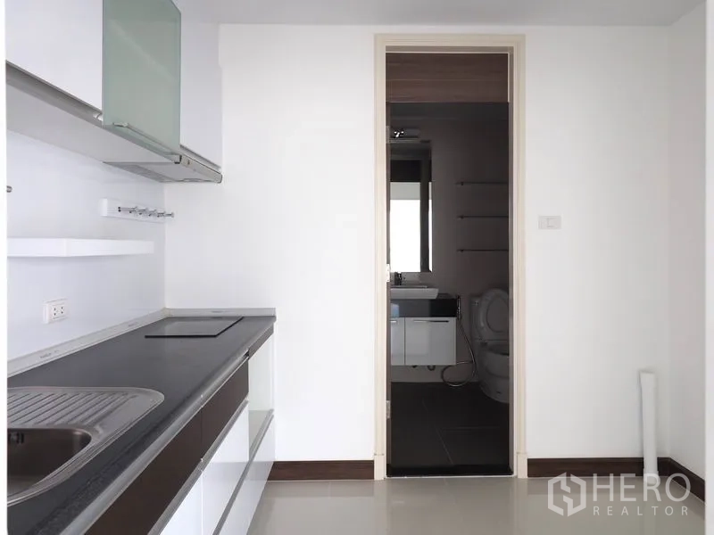 Condominium for rent in Yan Nawa, Bangkok - Minimal white kitchen with built-in hob, sink and cabinets, connected to a bathroom.