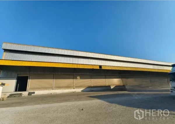 คลังสินค้า ให้เช่าในSamut Prakan - ด้านหน้าโกดังยาว หลังคาแถบสีเหลือง พร้อมประตูม้วนหลายช่อง ใต้ท้องฟ้าโปร่งใส