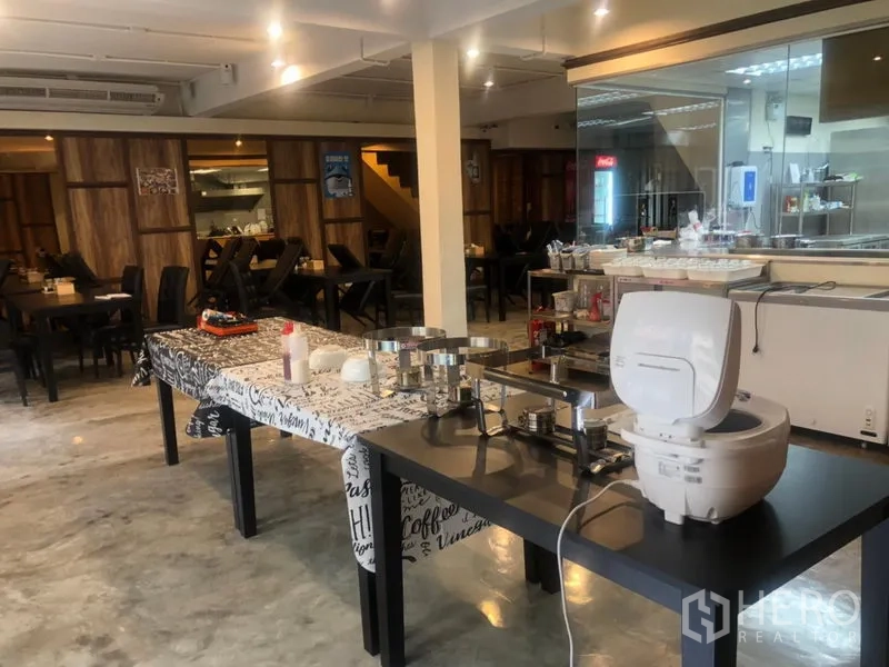 店屋 出租在Watthana, Bangkok - 大型餐厅楼层，摆放自助设备，玻璃后方为开放式厨房与多组餐桌。