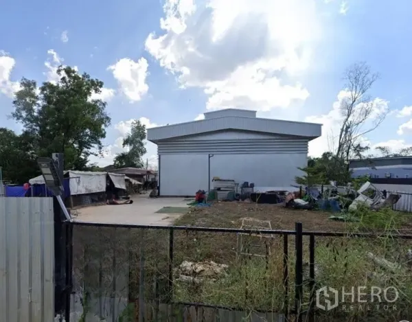 Warehouse for sale in Mueang Nonthaburi, Nonthaburi - Front view of gated property showing a large silver warehouse and open yard under a blue sky.