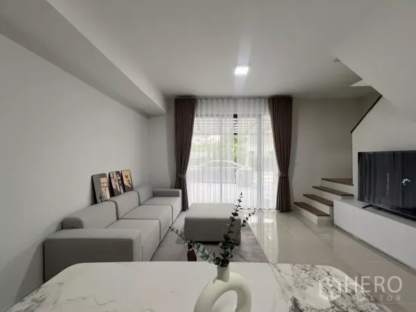 ทาวน์เฮาส์ ให้เช่าในBang Phli, Samut Prakan - ห้องนั่งเล่นสว่าง โซฟาสีเทาขนาดใหญ่ ม่านสูงจรดเพดาน ทีวีและบันได