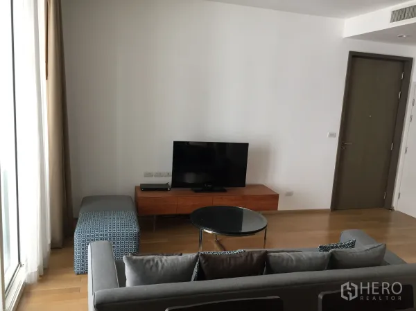 公寓 出租或出售在Watthana, Bangkok - 明亮客厅，灰色沙发、玻璃茶几、木质电视柜与落地窗帘。